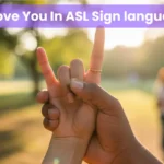 Person signing I Love You in ASL sign language outdoors in a sunny park with raised index and pinky